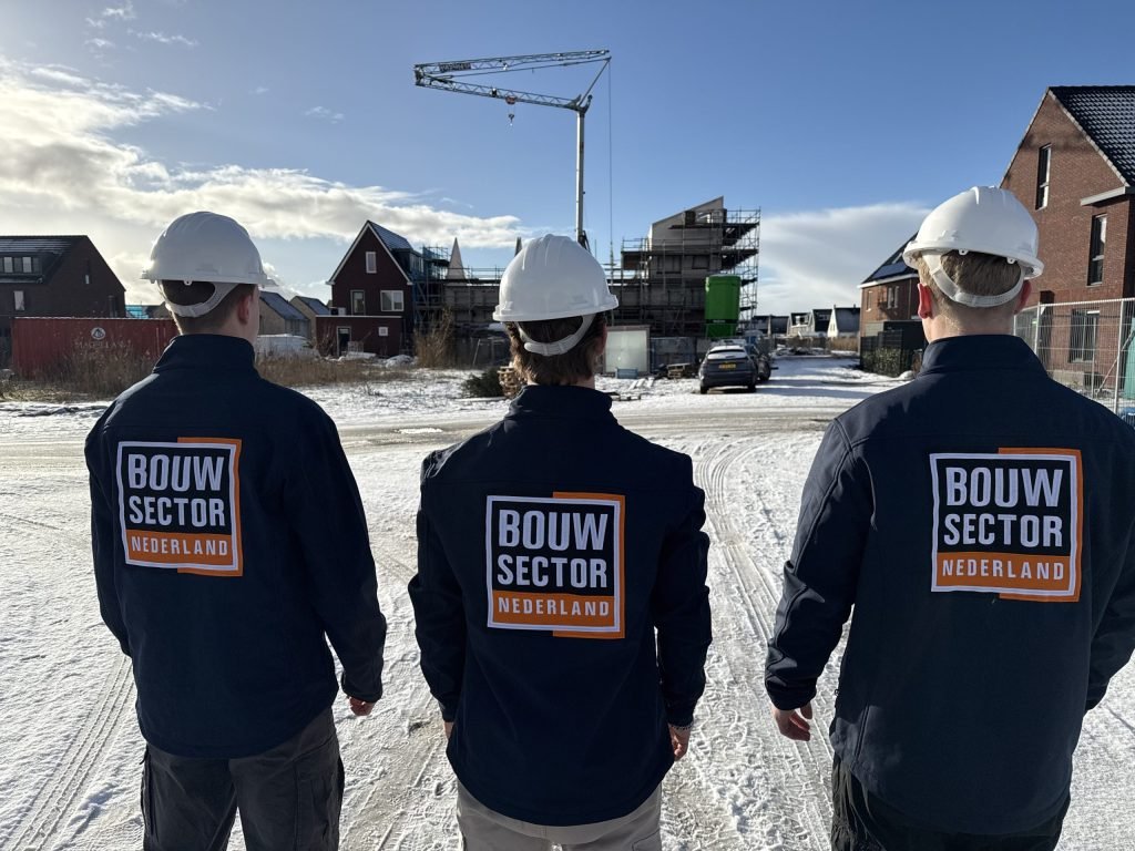 Drie teamleden in donkerblauwe werkjassen met logo kijken uit over een besneeuwde bouwplaats
